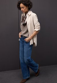 Chemise texturée beige clair portée sur un haut gris et un jean large bleu. Une écharpe sombre à motifs autour du cou et des bottines en daim marron.