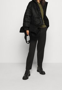 Osoba oblečená v černé péřové bundě, olivově zeleném topu, černých kalhotách a černých šněrovacích botách, držící složenou černou prošívanou tašku.