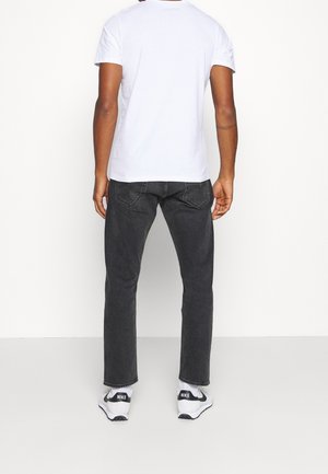 Mand iført en hvid t-shirt, sorte jeans og hvide Nike-sneakers, stående med ryggen mod en ensartet hvid baggrund.