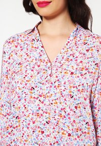 Blomstret bluse med hvid baggrund, prydet med et flerfarvet mønster af små blomster, V-hals og knapdetaljer.