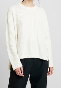 Maglione lavorato a coste di colore crema con scollatura rotonda, maniche lunghe e una patch del logo CK sull'orlo, abbinato a pantaloni scuri.