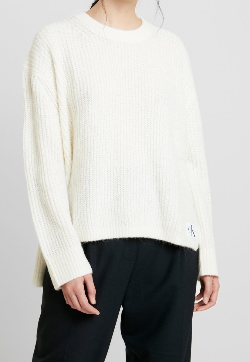 Maglione lavorato a coste di colore crema con scollatura rotonda, maniche lunghe e una patch del logo CK sull'orlo, abbinato a pantaloni scuri.