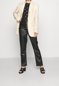 Cremfarbener Blazer über einem schwarzen gemusterten Oberteil, kombiniert mit hochtaillierten schwarzen Lederhosen und schwarzen klobigen Sandalen mit silbernen Hardware-Akzenten.