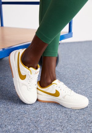 Jambes en leggings verts portant des baskets blanches avec le logo Nike jaune moutarde et des semelles en gomme, debout sur un tapis bleu près d'une étagère métallique bleue.
