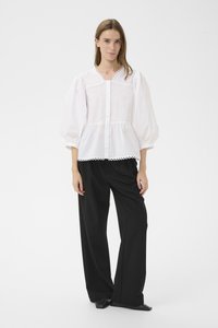 Hvid bluse med pufærmer, knappelukning foran og dekorativ kant ved halsudskæringen og kanten, kombineret med sorte bukser med vide ben og spidse sko.
