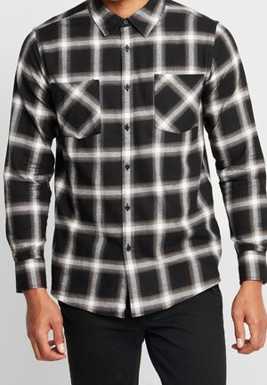 Schwarz-weiß kariertes Hemd mit Knöpfen und zwei Brusttaschen, getragen von einer Person vor einem schlichten Hintergrund.