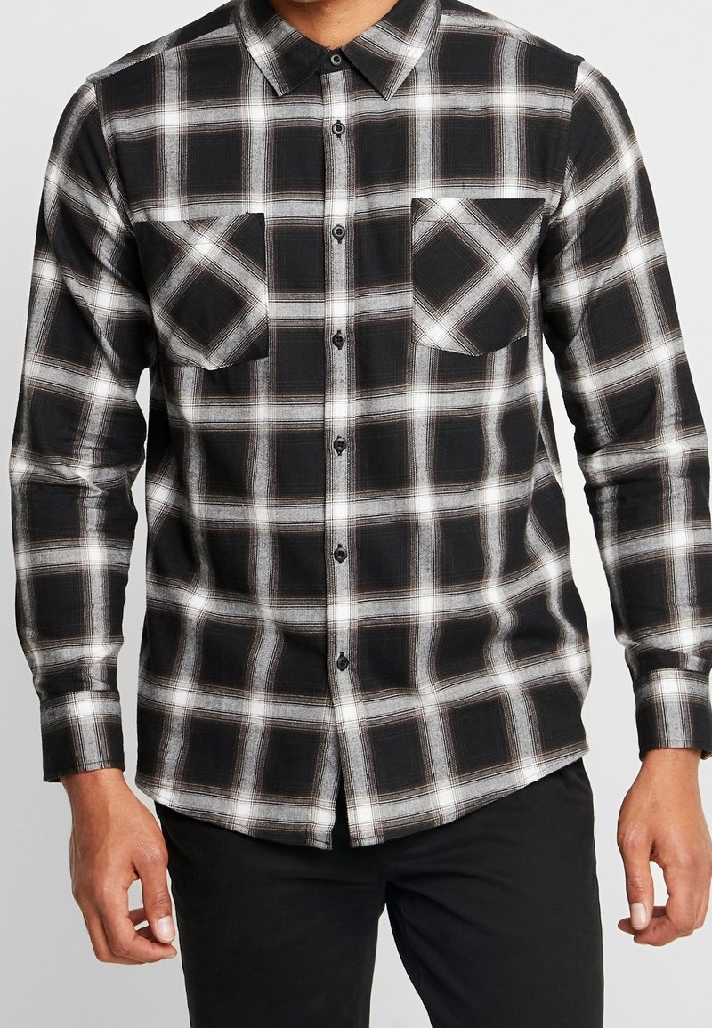 Chemise à carreaux noir et blanc à boutons, avec deux poches poitrine, portée par une personne se tenant devant un fond uni.