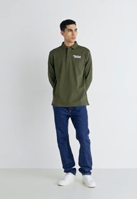 Zelena dolgi rokav polo majica s kragnico in sprednjim logotipom, v kombinaciji s temno modrimi kavbojkami in belimi supergami, stoji ob svetli ozadju.