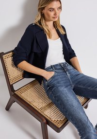 Chaqueta recortada azul marino con acentos de botones, camiseta blanca y jeans de un azul oscuro, sentado en una silla de mimbre con un marco de madera.