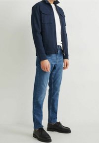 Chaqueta azul marino con cremallera y dos bolsillos en el pecho, usada sobre una camiseta blanca y unos jeans azules, combinada con zapatos negros de cordones.