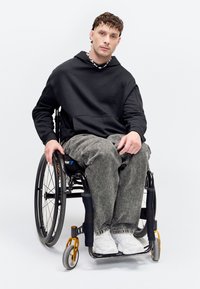 Jeune homme assis en fauteuil roulant, portant un sweat à capuche noir, un jean gris, des baskets blanches et un collier de perles sur un fond blanc uni.