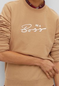 Sudadera beige con puños y dobladillo acanalados, con texto bordado en blanco "BE A Boss" en el frente. Tela suave y casual.