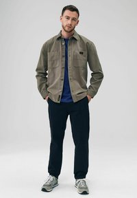Olijfgroene shirtjas met twee borstzakken, marineblauw t-shirt eronder, zwarte broek en grijze sneakers met witte accenten.