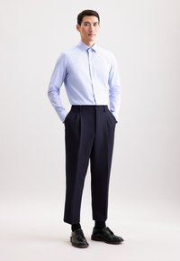 Lichtblauw, gestreept overhemd met een puntkraag, gecombineerd met marineblauwe geplooide broek. Zwarte nette schoenen maken de outfit compleet.