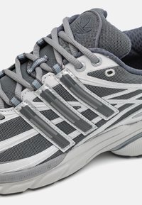 Sive atletske čevlje z mrežastim zgornjim delom, odsevnimi srebrnimi detajli in debele teksturirane podplati. Ima sive vezalke in tri vodoravne črte.