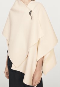 Crème-kleurige asymmetrische poncho met een metalen bladvormige broche, gedragen over zwarte kleding, die het bovenlichaam en de armen bedekt.