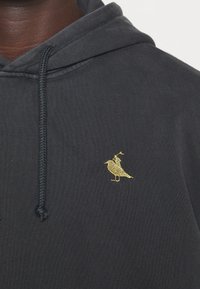 Sudadera con capucha de color gris oscuro con un logotipo de pájaro bordado en oro en el lado izquierdo del pecho. Tiene capucha con cordón y textura de tela suave.