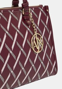 Borsa a mano bordeaux con un design a motivo che presenta il testo "Armani Exchange" e un charm a forma di cerchio dorato. Texture liscia con chiusura a zip.
