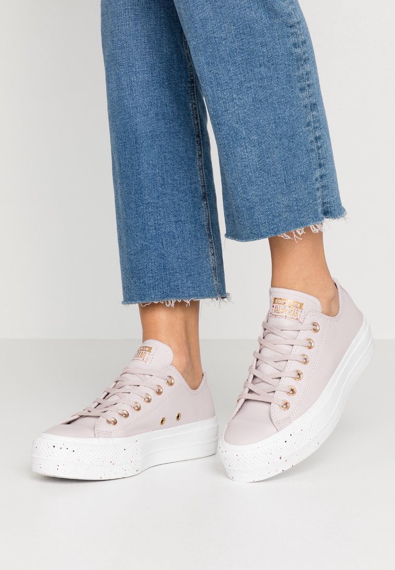 Zapatillas de lona rosa claro con una suela blanca gruesa, que cuentan con ojales dorados y una puntera texturizada. El diseño incluye un logo en la lengüeta.