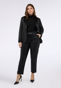 Kvinna som står självsäkert klädd i en glänsande svart kavaj, matchande högmidjade byxor, svart polotröja och svarta klackar, mot en vit bakgrund.