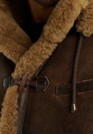 Bruine schapenvachtjas met een tan, fluffy kraag; heeft een houten toggle-sluiting en leren accenten. Getextureerde buitenkant, warm ontwerp.