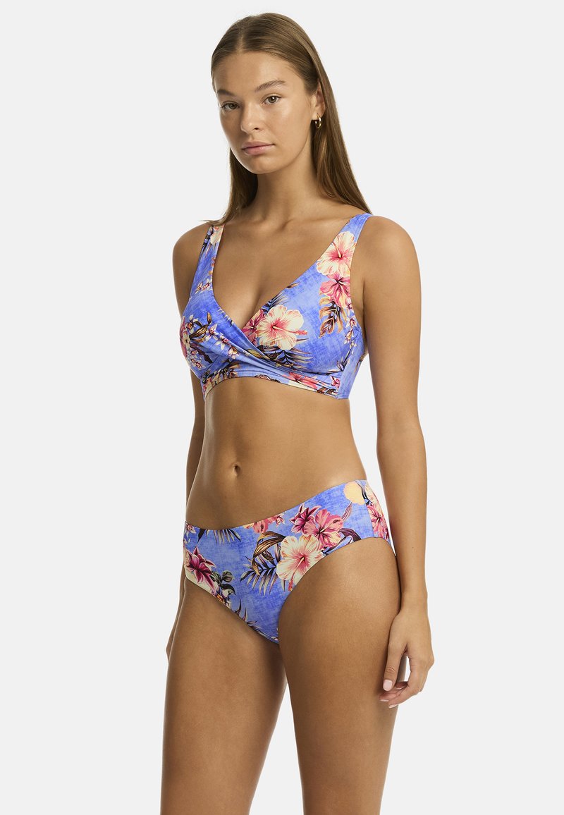 Een jonge vrouw poseert zelfverzekerd in een blauwe bloemenbikini, versierd met roze en gele bloemen, met zachte golven in haar haar, tegen een effen achtergrond.