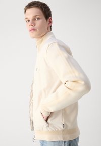 Ljusbeige zip-jacka med ribbad krage och muddar, med sidofickor och en jämn textur. Minimalistisk design, inga framträdande mönster.