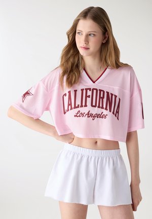 Giovane donna con capelli lunghi indossa una maglietta rosa corta in rete con la scritta "California Los Angeles" e pantaloncini a vita alta bianchi con elastico, in posa con una mano sui fianchi.