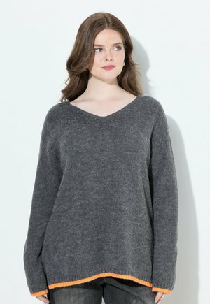Femme portant un pull gris ample à col en V avec des bordures orange aux poignets et à l'ourlet, associé à un jean noir déchiré, sur un fond uni.