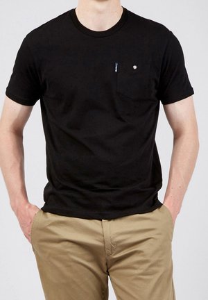 Homme portant un t-shirt noir à manches courtes avec une petite poche poitrine et un pantalon beige, les mains dans les poches.