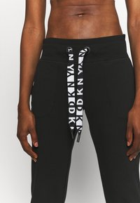 Svarta träningbyxor med två framfickor, har en ribbad midja och snörning med ett vitt logomönster. Slät textur.