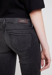 Vaqueros de mezclilla negra con un diseño de cinco bolsillos, un ligero desgastado y un parche de cuero con la marca en la parte posterior de la cintura.