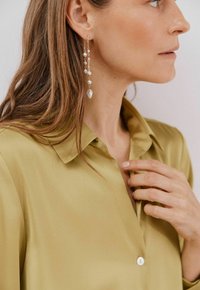 Blusa in satin verde oliva con colletto e apertura con bottoni, abbinata a orecchini pendenti con perle e accenti dorati.