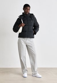 Giubbino trapuntato nero con colletto alto e chiusura a zip, abbinato a pantaloni larghi grigi chiari. Sneakers bianche completano l'outfit.