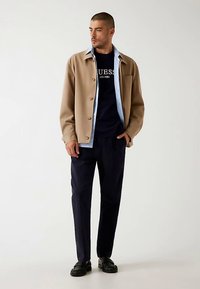 Giacca beige con bottoni su una felpa blu navy con logo bianco, abbinata a pantaloni scuri e scarpe nere. Tessuti lisci e design casual.