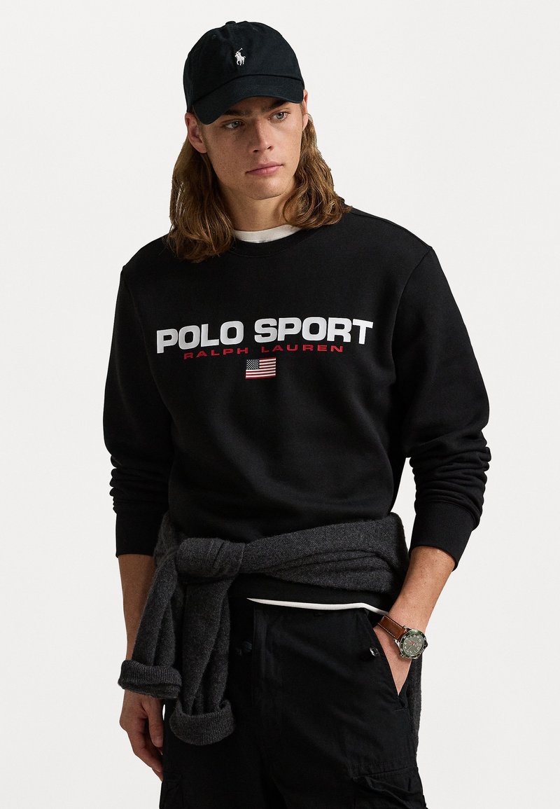 Hanorac negru cu textul "POLO SPORT" în alb și roșu, având un steag american. Purtați cu un pulover gri închis legat la brâu.