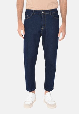 Pantaloni in denim blu scuro con una vestibilità leggermente ampia, cuciture visibili e tasche frontali. Indossati con sneakers bianche.
