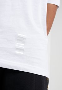 Bílé bavlněné tričko s hladkou texturou a svislou řadou reflexních stříbrných pruhů na levé dolní straně.