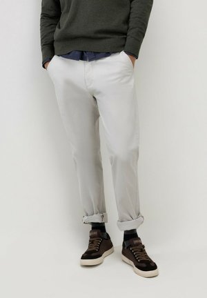 Hombre con pantalones gris claro remangados, zapatillas marrón oscuro, calcetines negros, suéter verde oscuro y camisa azul marino con las manos en los bolsillos.