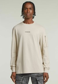Jasnobeżowa bluzka z długim rękawem o heksagonalnej teksturze, z logo na przodzie i boczną kieszenią na zamek. Miękka tkanina i luźny krój.