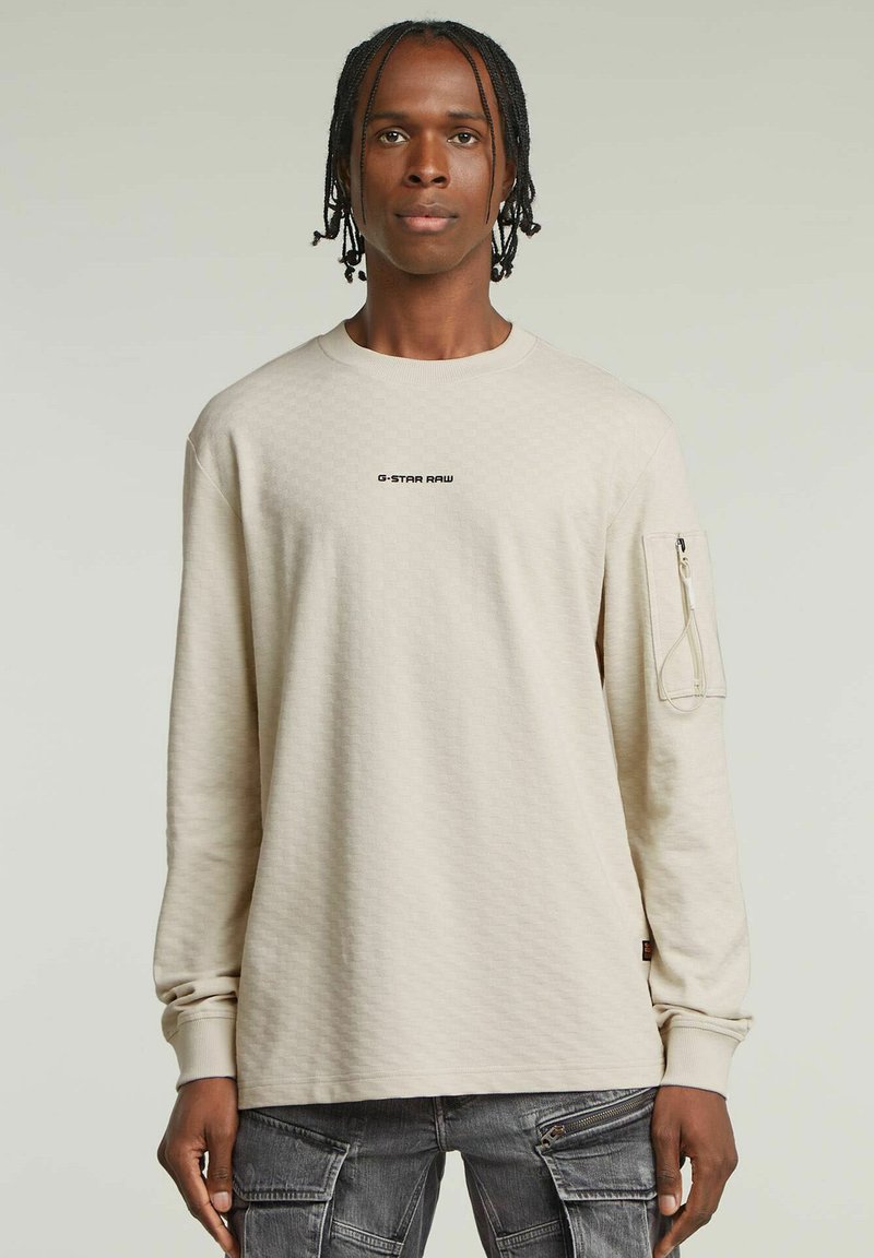 Jasnobeżowa bluzka z długim rękawem o heksagonalnej teksturze, z logo na przodzie i boczną kieszenią na zamek. Miękka tkanina i luźny krój.