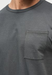 Κοντινό πλάνο ενός ατόμου που φοράει σκούρο γκρι μπλουζάκι με στρογγυλή λαιμόκοψη και ορατή τσέπη στο στήθος στην αριστερή πλευρά.