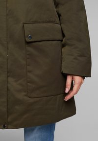 Chaqueta aislante verde oliva con un gran bolsillo plano y detalle de botón de presión. Textura suave, de longitud media y ajuste casual.