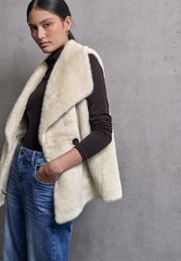 Gilet in pelliccia sintetica color crema con colletto scialle e chiusura con bottone, indossato sopra un top marrone e jeans blu, caratterizzato da una vestibilità comoda e una consistenza morbida.