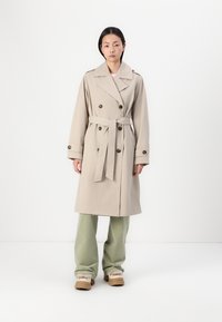 Beige trenchcoat med dobbelknapplet front, store knapper og bælte. Har brede revers, lange ærmer og en glat stoftekstur.
