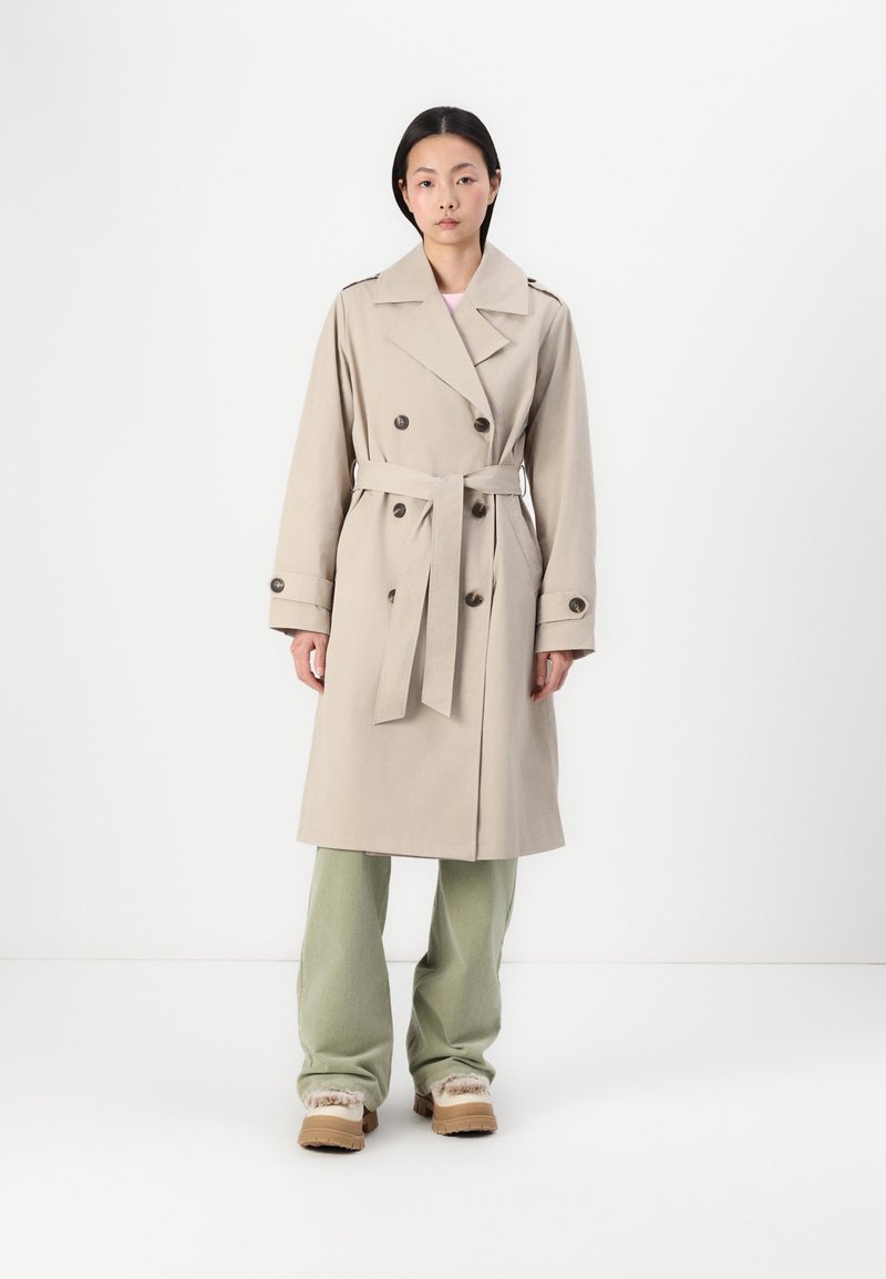 Beige trenchcoat med dobbelknapplet front, store knapper og bælte. Har brede revers, lange ærmer og en glat stoftekstur.