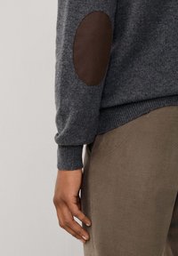 Pull en maille grise avec des coudières en cuir marron, des poignets et un ourlet côtelés, associé à un pantalon marron clair. Présente des textures douces et des lignes épurées.