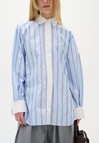 Šviesiai mėlynas marškinėliai su balta apykakle ir rankogaliais. Yra vertikalūs mėlyni dryžiai, mygtukų užsegimas priekyje ir laisvas siluetas. Pagaminti iš medvilnės audinio.