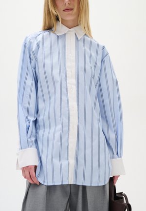 Camicia a righe azzurre con colletto e polsini bianchi. Presenta righe verticali blu, chiusura con bottoni sul davanti e vestibilità ampia. Tessuto in cotone.