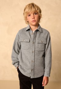 Veste-chemise grise à motif chevrons avec deux poches poitrine, boutonnée sur le devant, au tissu doux, associée à un pantalon en velours côtelé foncé.